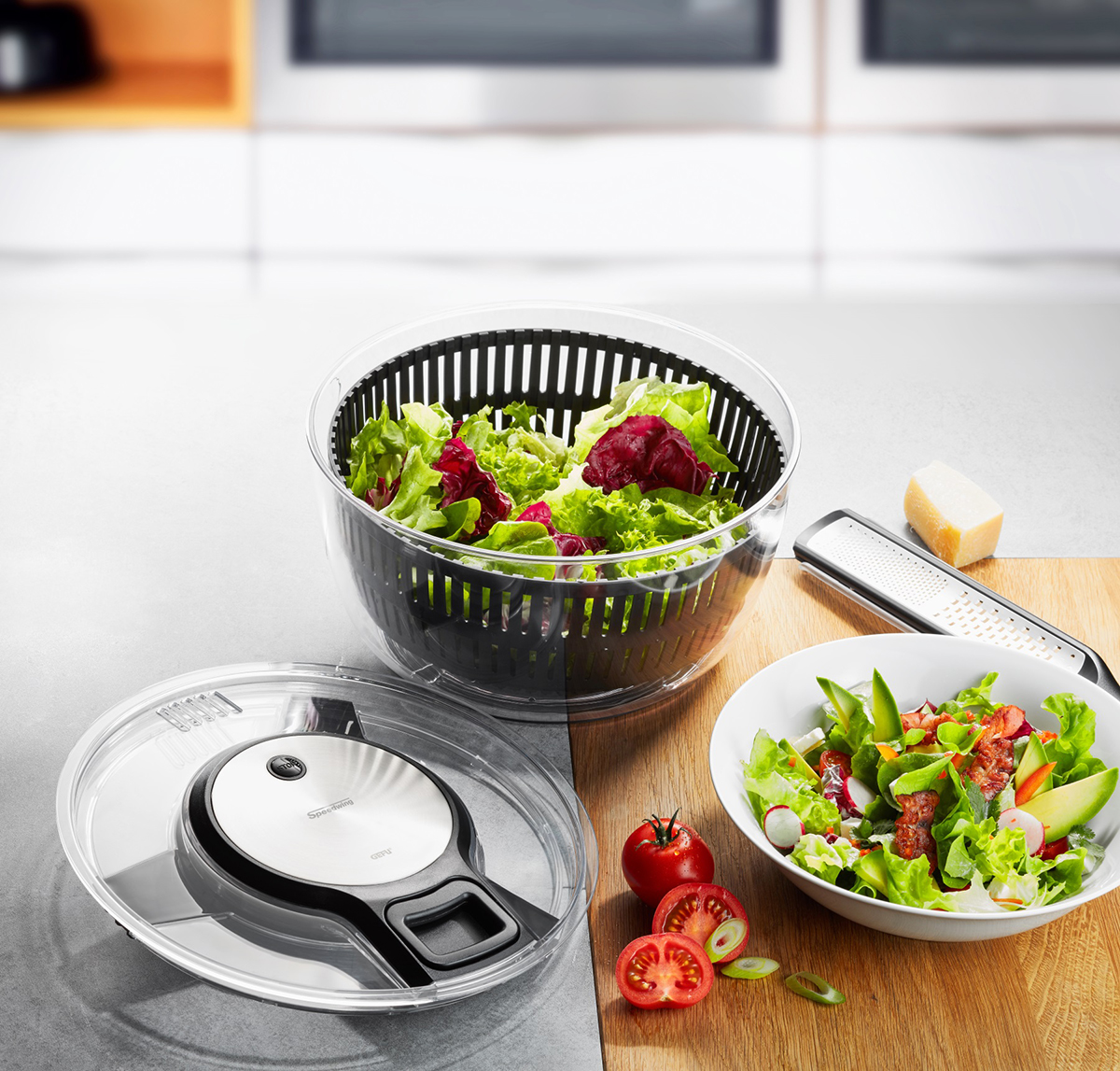 Salatschleuder mit frischem Gemüse und Küchenutensilien auf dem Tisch Eine Salatschleuder voller frischem Salat neben einer Schüssel mit gemischtem Salat, frischen Tomaten und einer Reibe auf einem Küchentisch