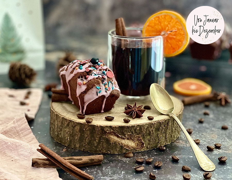 Kleine Kaffee-Cakes mit Glühweinguss Ein Glas Glühwein mit Orangenscheibe neben einem Schokoladenkuchenstück mit rosa Zuckerguss, dekoriert mit Zimtstangen und Kaffeebohnen auf einem rustikalen Holztisch