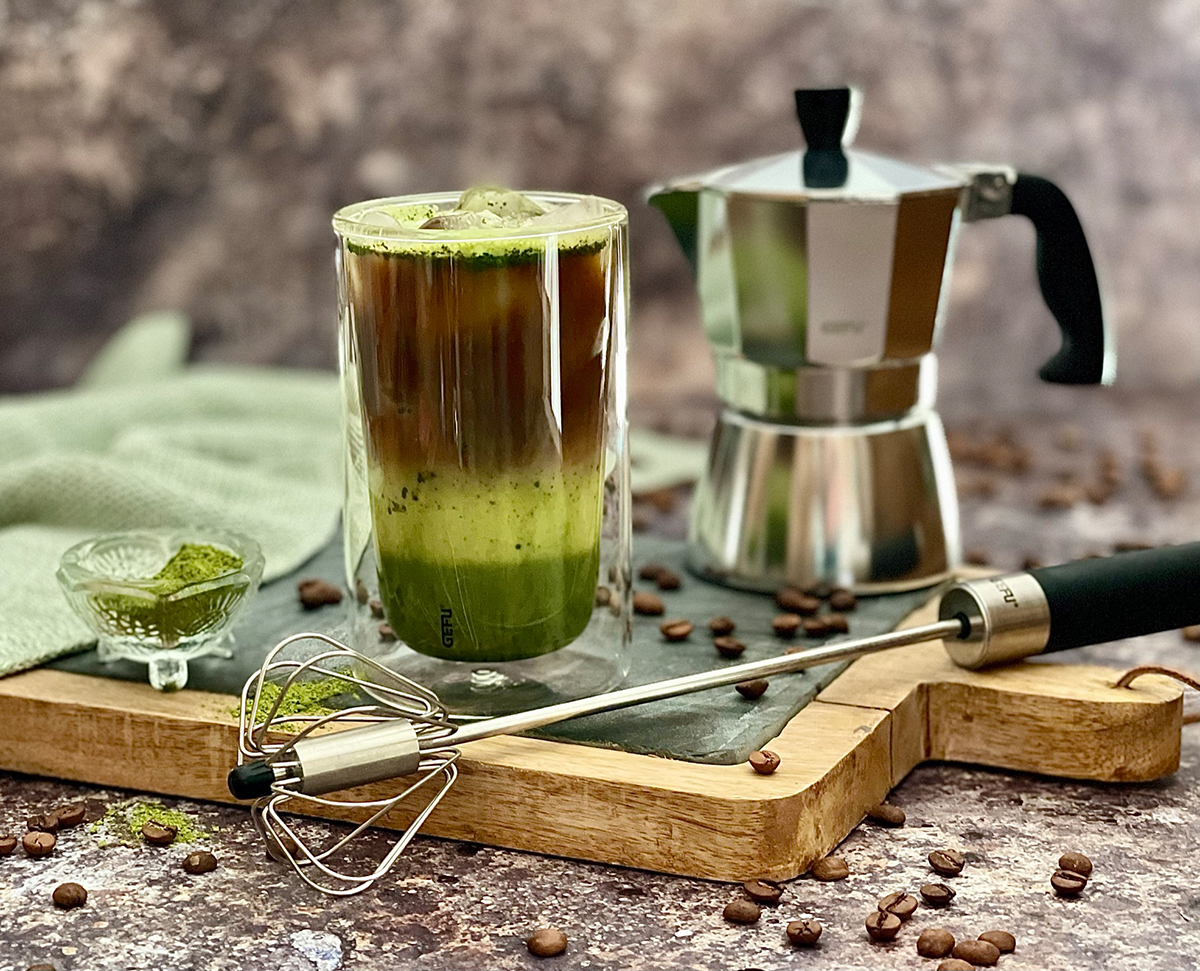 Matcha-Eiskaffee mit Kaffeemaschine und Schneebesen Ein Glas Matcha-Eiskaffee steht auf einem Tablett neben einer Kaffeekanne und einem Schneebesen, umgeben von Kaffeebohnen und Matchapulver.
