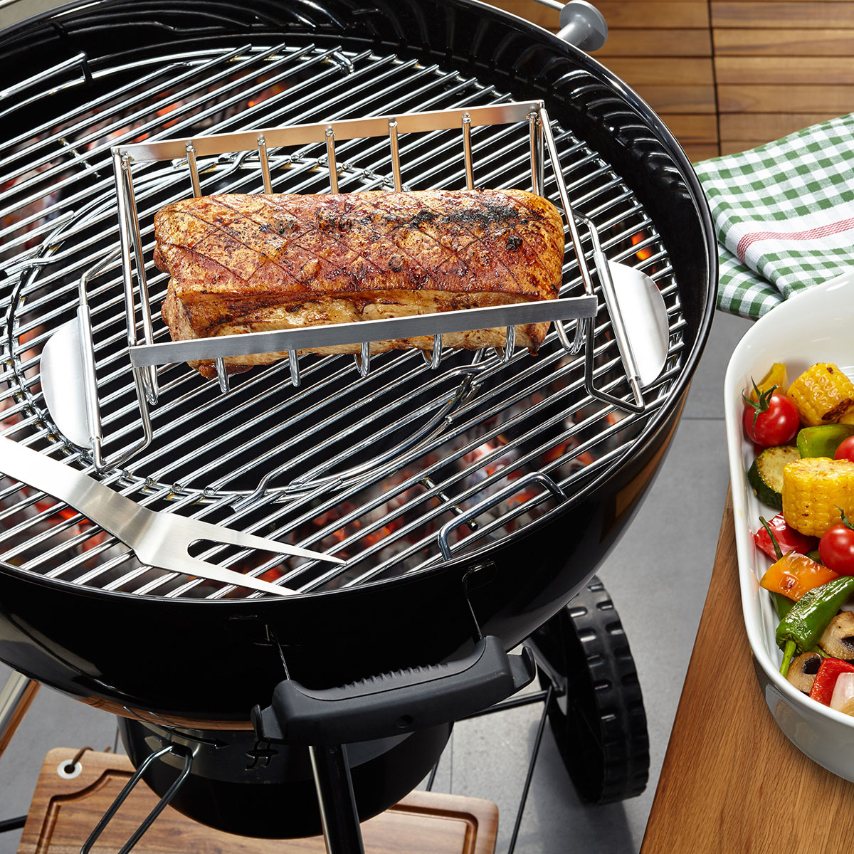 Ein Rundgrill mit Fleisch in einem Halter, daneben eine Schale mit Gemüse.