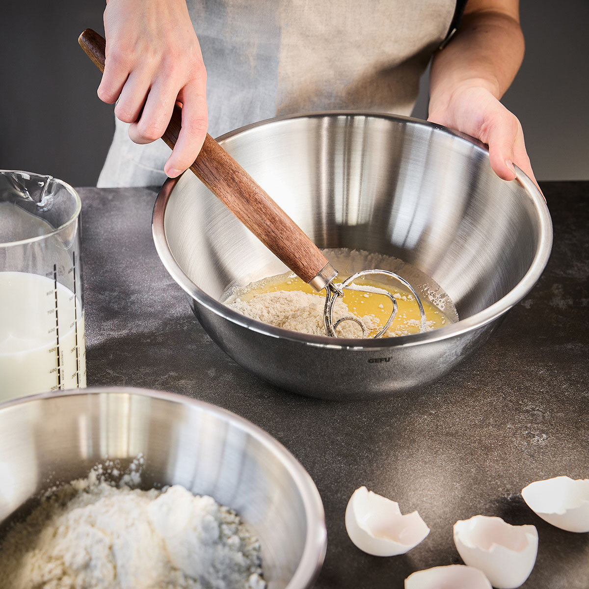 Teigmischung in Edelstahlschüssel mit Schneebesen Person rührt Teig in einer Edelstahlschüssel, umgeben von Zutaten wie Milch, Mehl und Eierschalen.