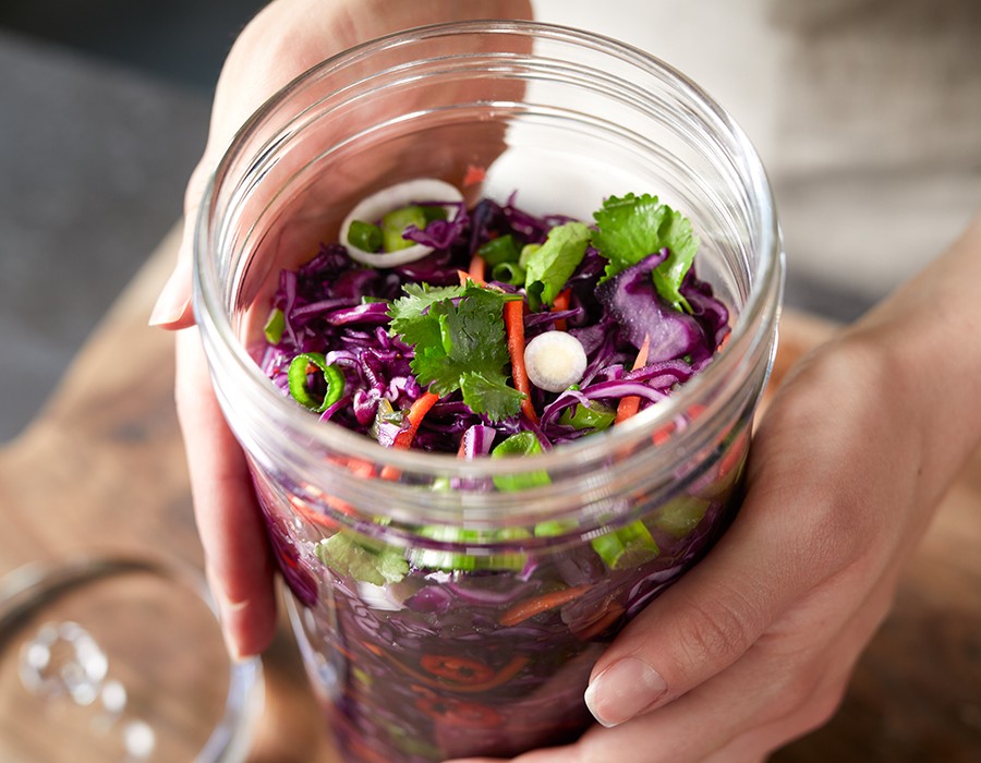 Rotes Sauerkraut mit Möhren, Frühlingszwiebeln, Koriander und Chili Nahaufnahme eines Glases mit selbstgemachtem Kimchi, gefüllt mit frischem Gemüse wie roten Kohl, Karotten und Koriander. Zwei Hände halten das Glas auf einem Holztisch.