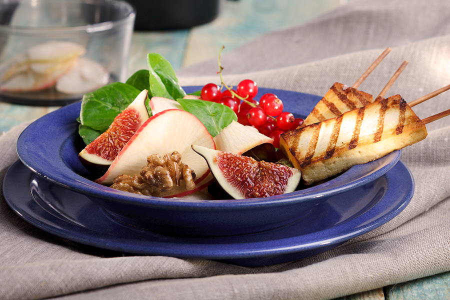 Apfel-Feigen-Salat mit Mangovinaigrette und gegrillten Tofusticks Ein blauer Teller mit frischer Salatplatte, die Feigen, rote Trauben, Walnüsse und gegrillte Käse-Spieße enthält, auf einem Leinentuch serviert.