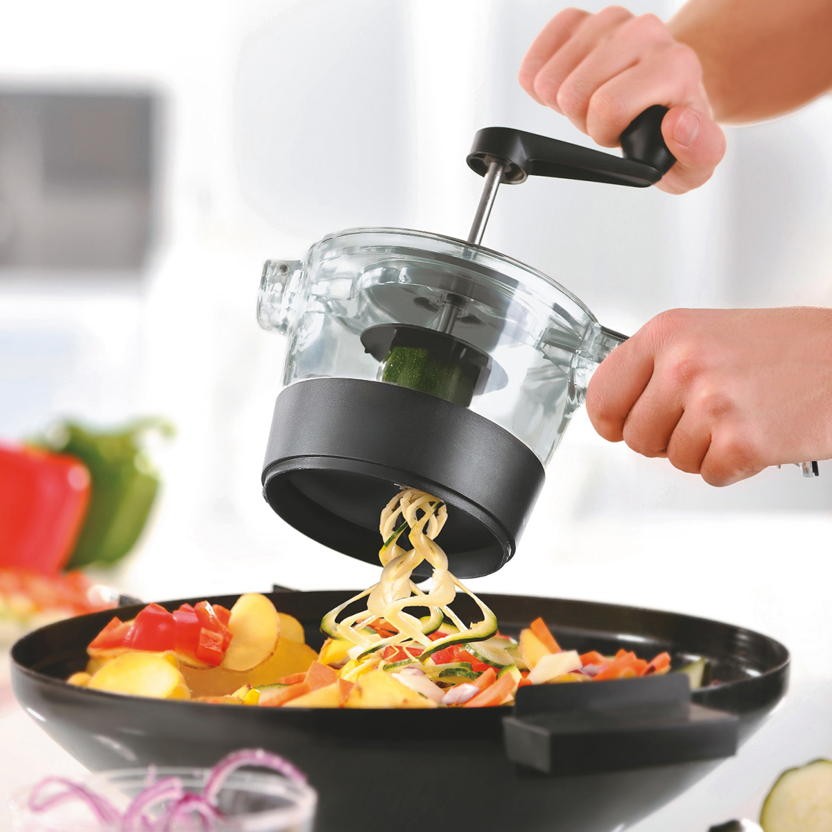 Handbetriebenes Küchengerät zur Herstellung von Gemüsespaghetti in einer Pfanne Person benutzt ein handbetriebenes Küchengerät, um Zucchini in Spiralen zu schneiden, die in eine Pfanne mit Gemüse fallen.