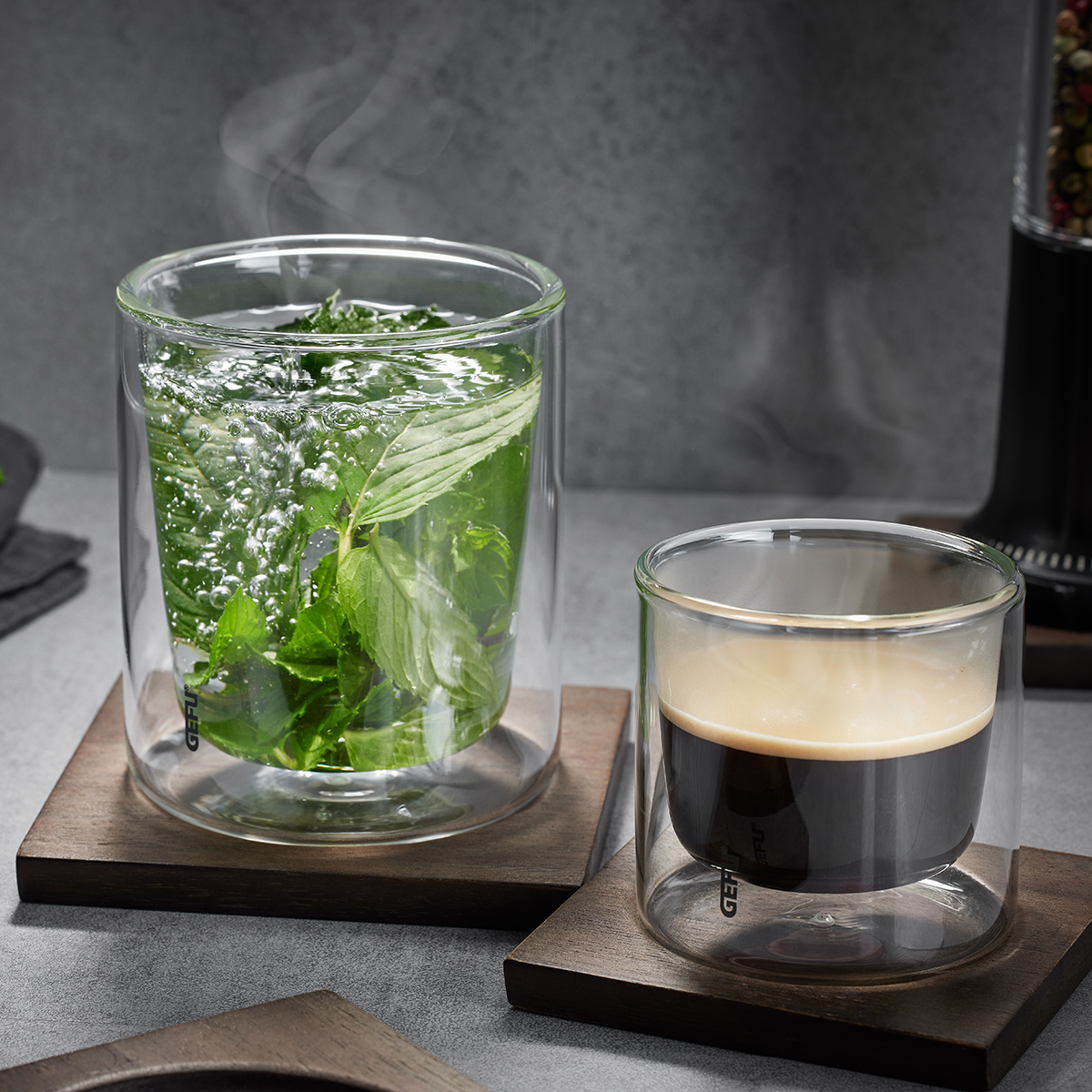 Doppelwandige Teegläser mit frischem Kräutertee und Espresso auf Holzuntersetzern Zwei doppelwandige Gläser auf Holzuntersetzern mit frischem Kräutertee und heißem Espresso, wobei Dampf aufsteigt.