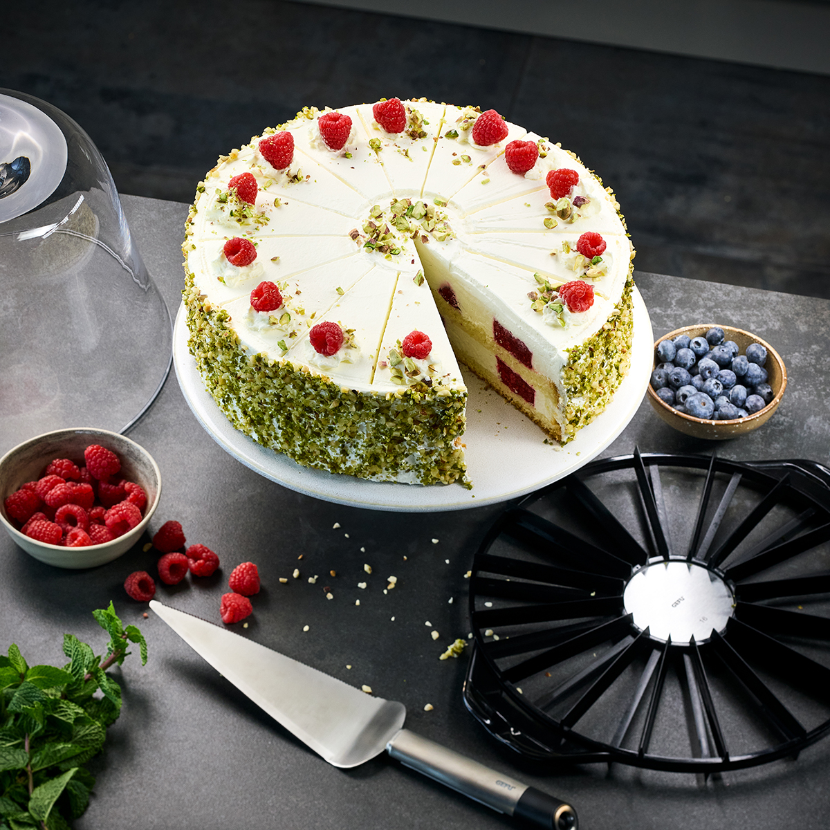 Eine angeschnittene Himbeer-Pistazien-Torte auf einem Tisch, umgeben von Schalen mit frischen Himbeeren und Blaubeeren sowie einem Messer