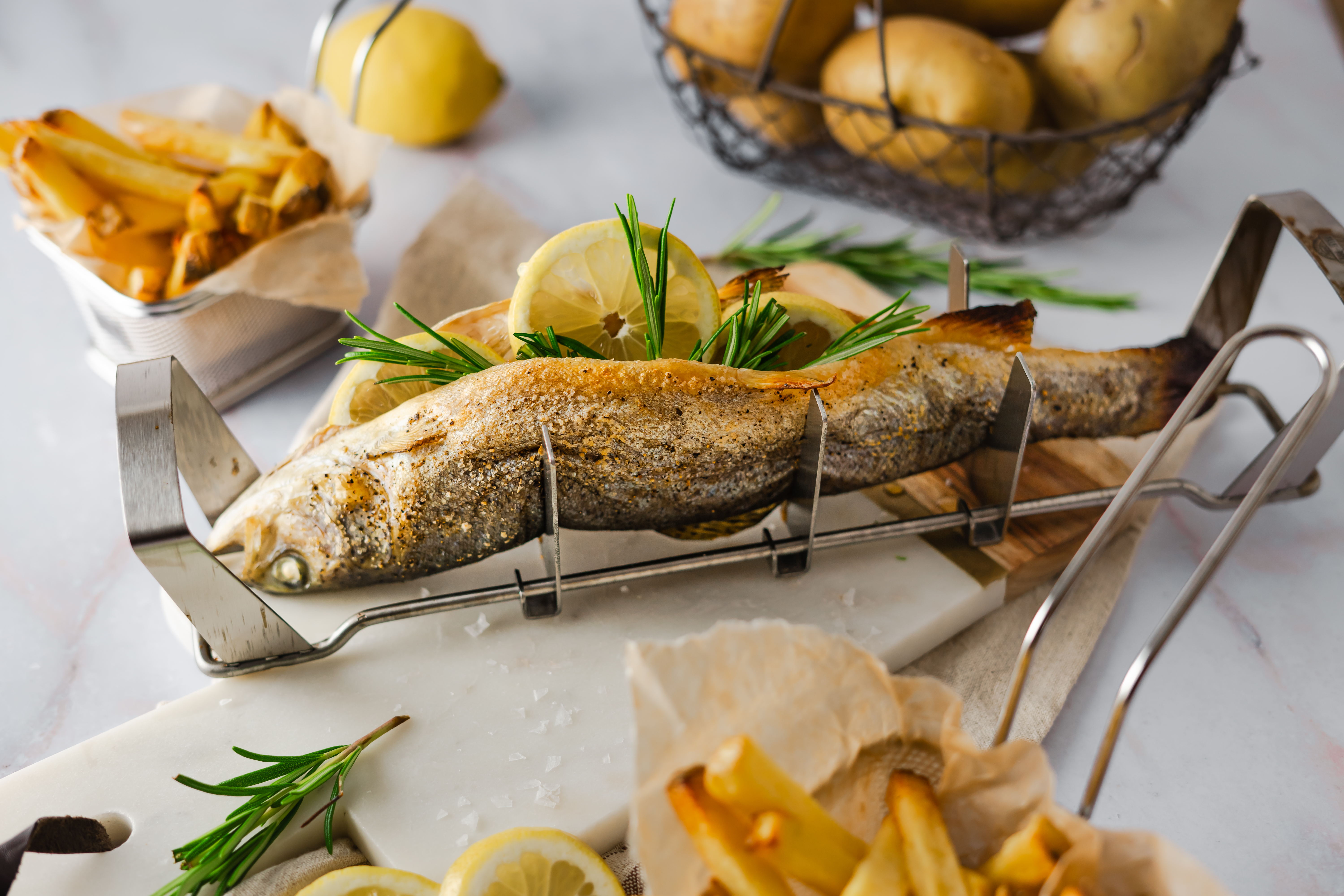 Rezept für gegrillte Forelle mit Zitrone und Rosmarin Ein gegrillter Fisch auf einem Metallhalter, garniert mit Zitronenscheiben und Rosmarin, umgeben von Pommes Frites und Kartoffeln