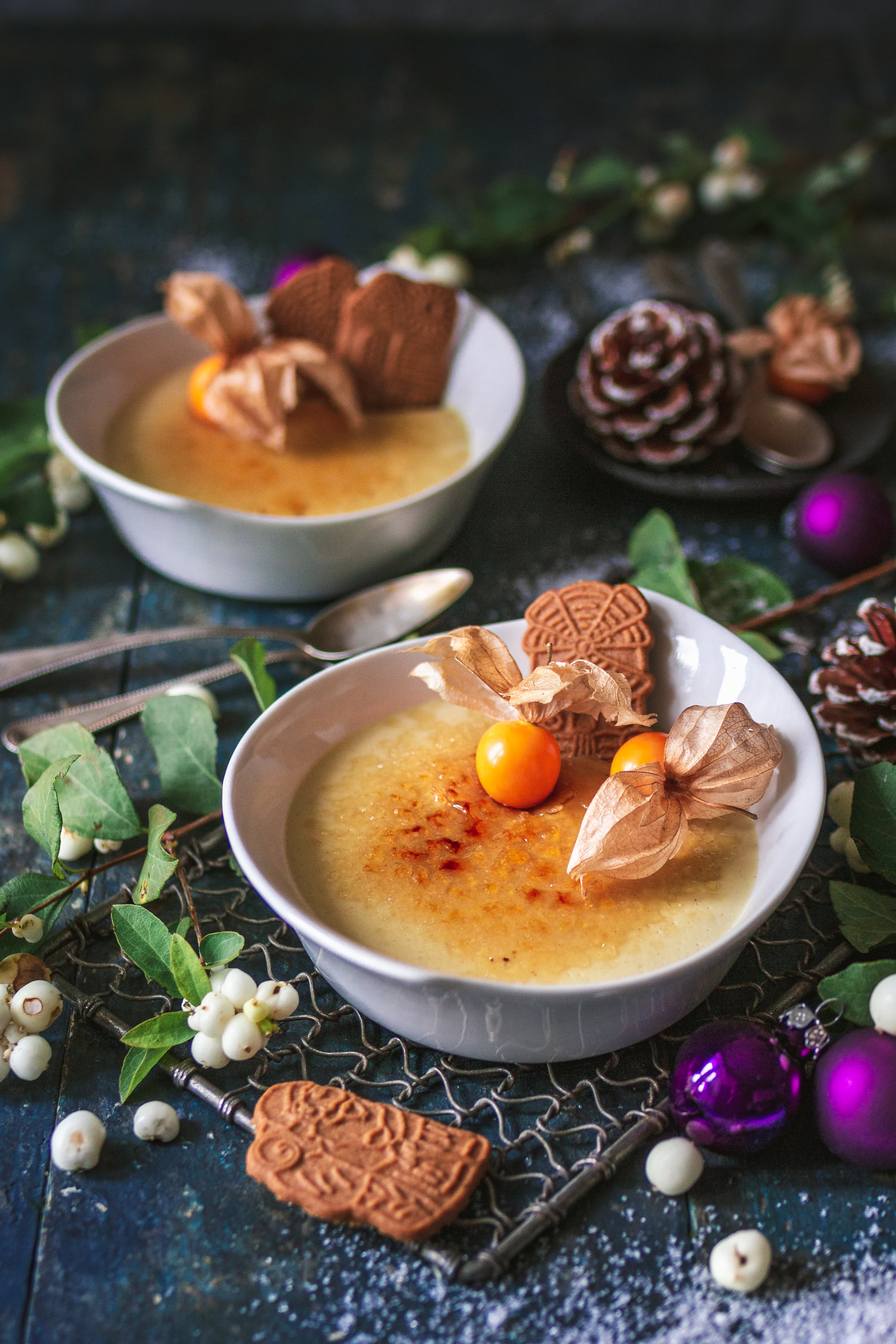 Creme Brulee Eine Schale Crème Brûlée, garniert mit Physalis und Spekulatius-Keks, umgeben von Tannenzapfen und Weihnachtsschmuck auf einem Tisch.