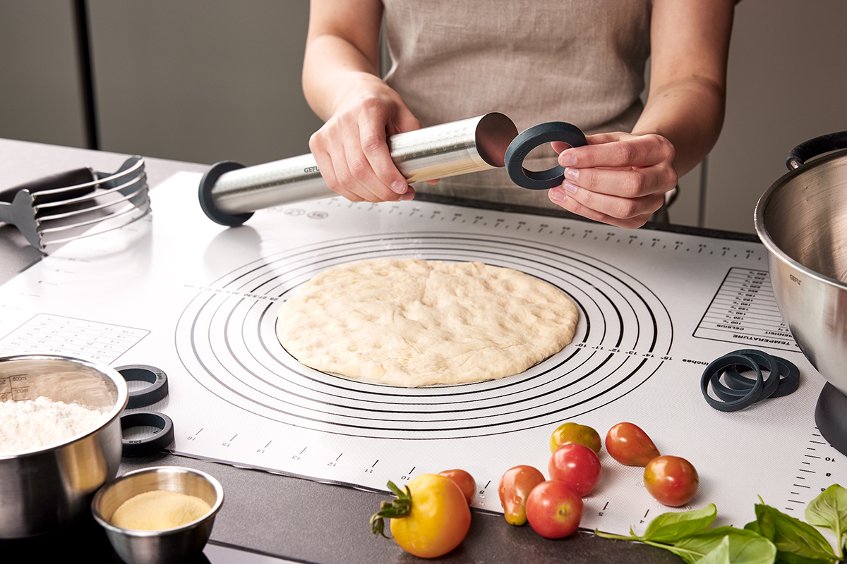 Teigzubereitung mit verstellbarem Nudelholz auf Silikonmatte Person passt ein verstellbares Nudelholz an, um Teig auf einer Silikonmatte auszurollen. Daneben liegen Mehl und frische Tomaten.