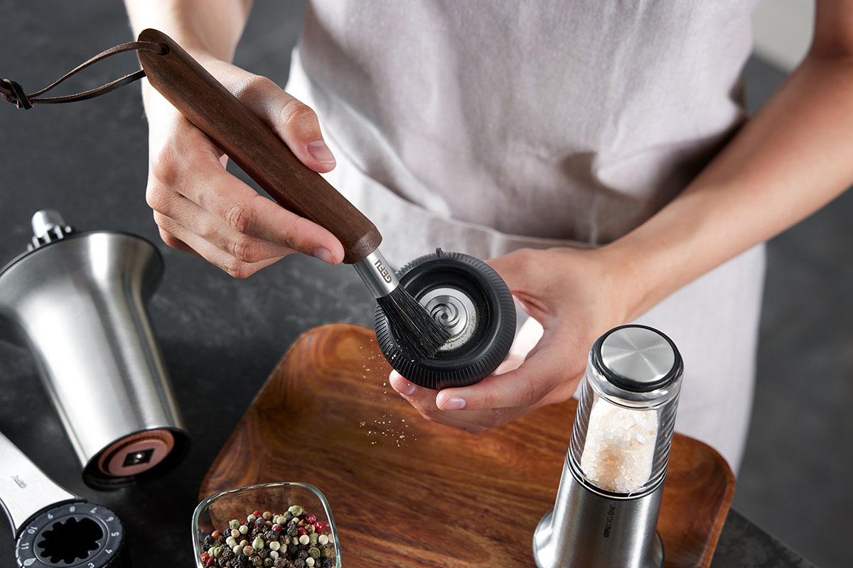 Praktische Reinigung eines Salz- und Pfeffermühlen-Sets mit Bürste Person reinigt das Innere einer Salz- und Pfeffermühle mit einer Bürste auf einer Holzoberfläche, mit Pfefferkörnern im Hintergrund