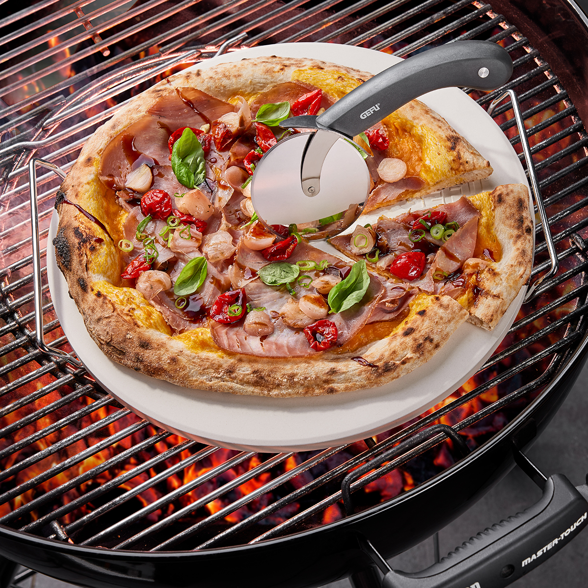 Frische Pizza auf Grill mit Pizzaschneider Pizza mit Schinken, Tomaten und Basilikum auf einem Grill, geschnitten mit Pizzaschneider
