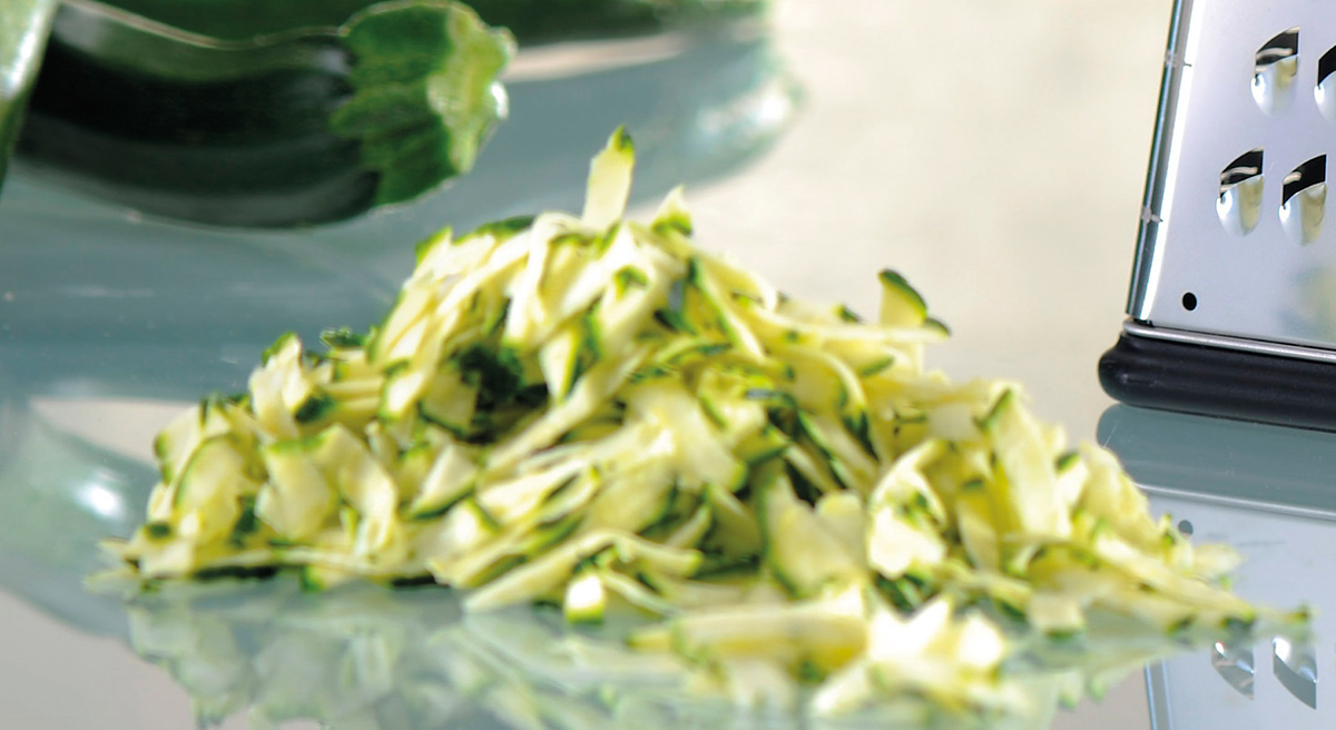 Frisch geriebene Zucchini neben Reibe auf Küchentisch Nahaufnahme von geriebener Zucchini auf einem Tisch mit einer Reibe im Hintergrund