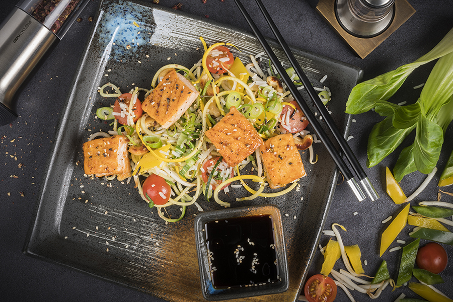 Asiatische Gemüse-Reis-Pfanne mit Lachswürfeln und Szechuanpfeffer Ein Teller mit asiatischer Nudelpfanne, Lachs und frischem Gemüse, garniert mit Sesam, serviert mit Sojasoße.