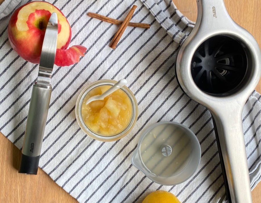 Apfelmus Ein halber Apfel, ein Schäler, ein Glas Apfelmus, Zimtstangen, ein Messbecher und eine Zitruspresse liegen auf einem gestreiften Küchentuch.