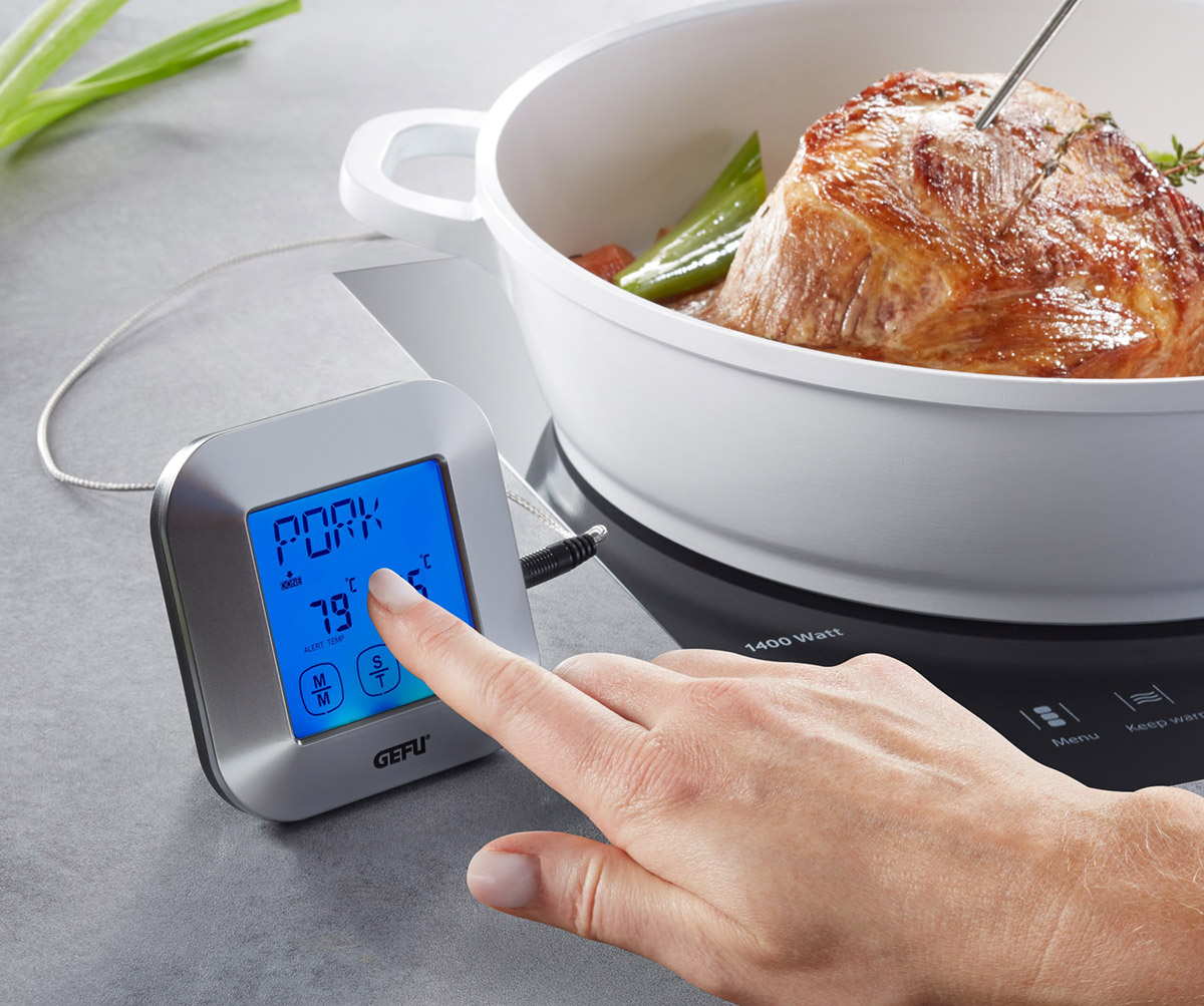 Digitales Fleischthermometer bei der Messung der Kerntemperatur von Schweinebraten Hand bedient ein digitales Fleischthermometer neben einem Topf mit Schweinebraten auf einem Herd