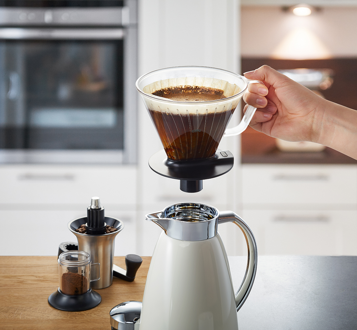 Handgebrühter Kaffee mit modernem Filterhalter in der Küche Eine Person verwendet einen Handkaffee-Filterhalter, um frischen Kaffee in eine Kanne zu gießen. Im Hintergrund ist eine moderne Küche zu sehen.