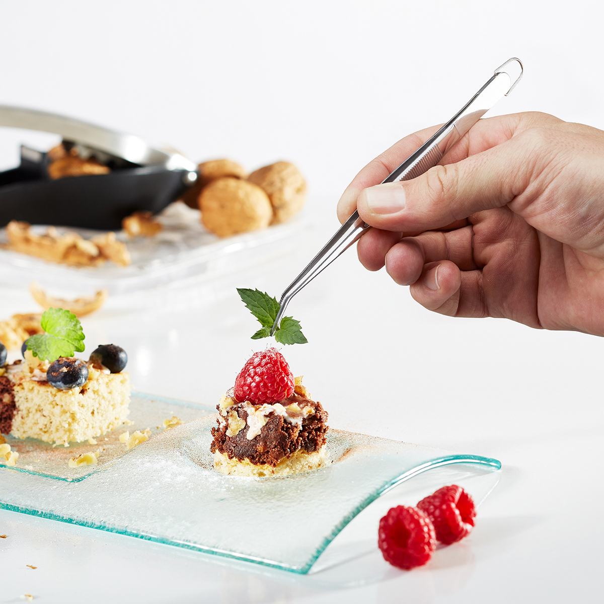 Naheaufnahme einer Hand, die ein Minzblatt mit einer Pinzette auf ein kunstvoll angerichtetes Dessert mit Himbeere legt