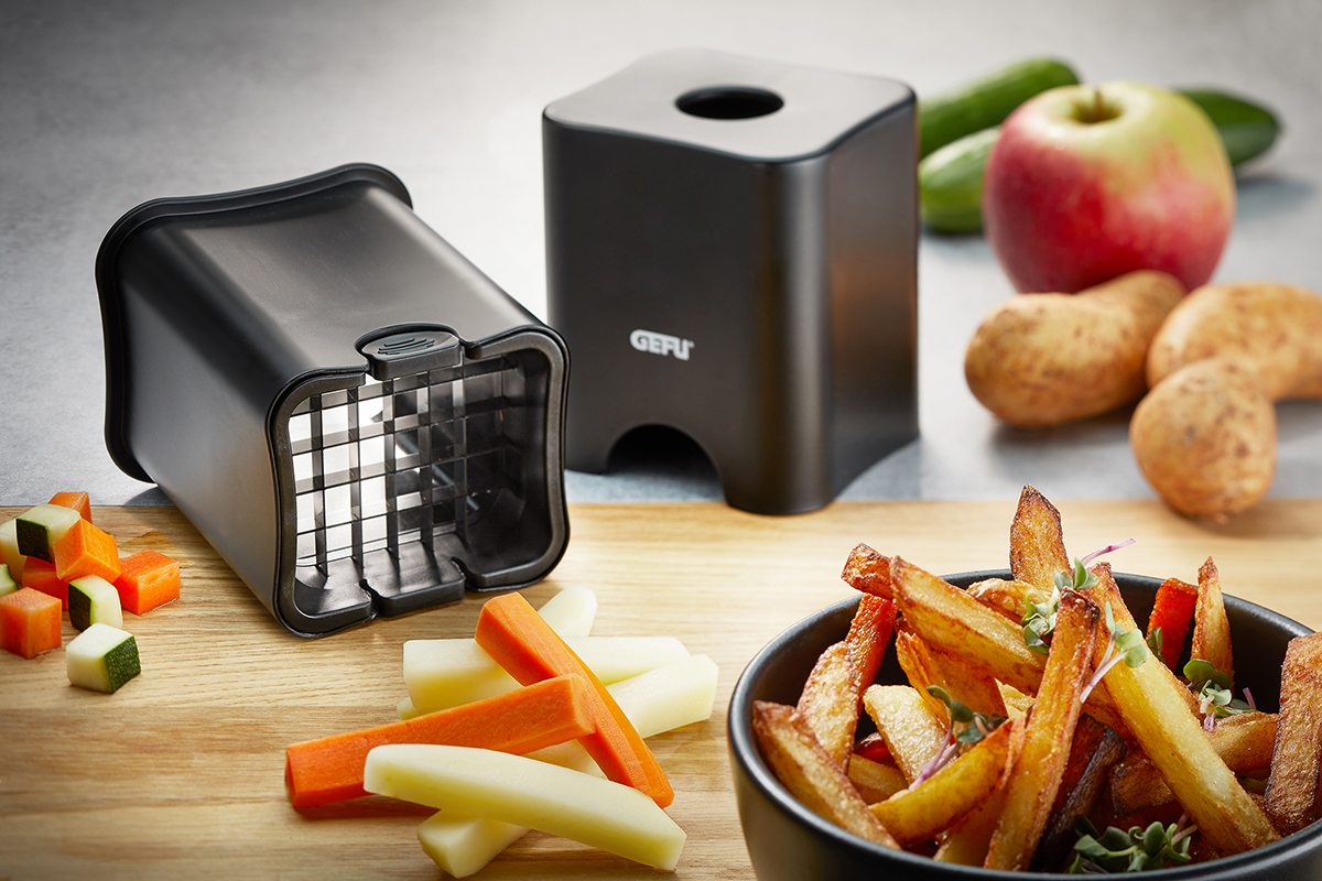 Innovativer Gemüseschneider für gleichmäßige Pommes und Gemüsesticks Schwarzer Gemüseschneider mit Würfeln und Pommes auf einem Tisch, umgeben von Kartoffeln, Karotten und einem Apfel