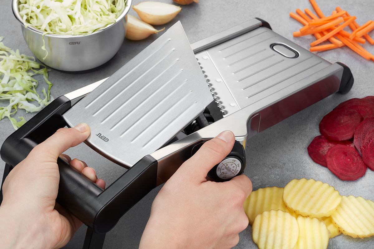 Hochwertige Gemüsereibe für gleichmäßige Scheiben Person verwendet eine Gemüsereibe, um verschiedene Gemüsesorten wie Karotten, Rote Beete und Kartoffeln in gleichmäßige Scheiben zu schneiden. Ein Topf mit gehobeltem Kohl ist ebenfalls sichtbar.