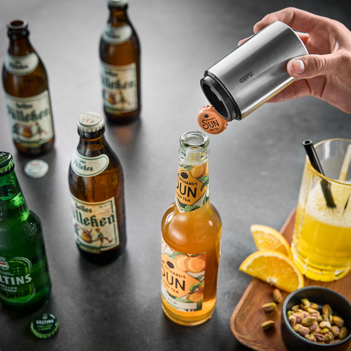 Hand benutzt einen Edelstahl-Flaschenöffner, um eine Flasche Eistee zu öffnen, umgeben von anderen Bierflaschen und einer Orangenscheibe auf dem Tisch