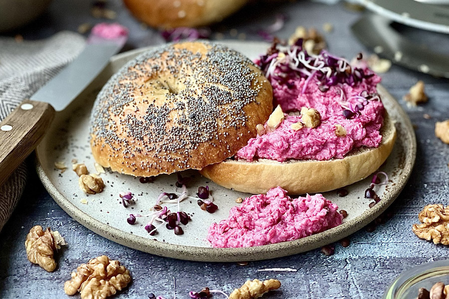 Bagels mit Rote Beete Aufstrich und Sprossen Ein Teller mit einem Mohnbrötchen, gefüllt mit Rote-Bete-Aufstrich und Walnüssen, dekoriert mit Sprossen.
