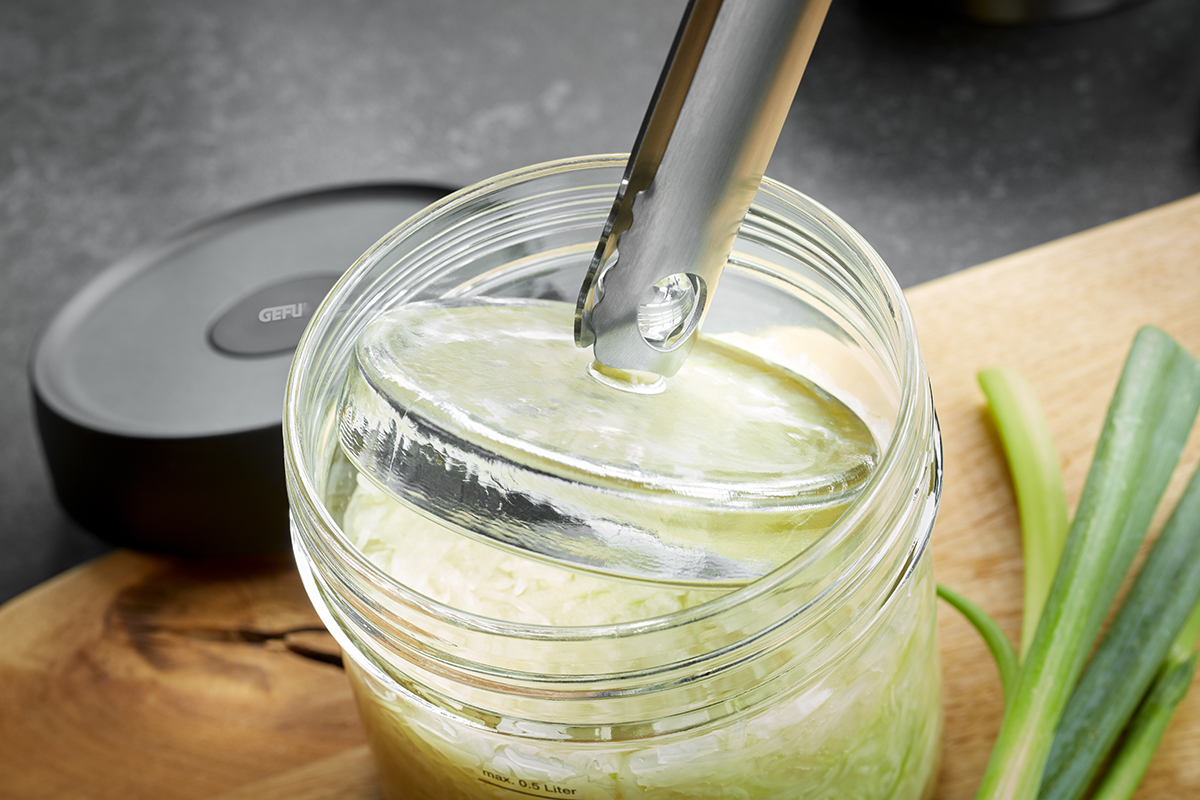 Ein Glasgefäß, gefüllt mit Sauerkraut, wird mit einer Edelstahlzange geöffnet. Auf einem Holzbrett liegt frischer Lauch.