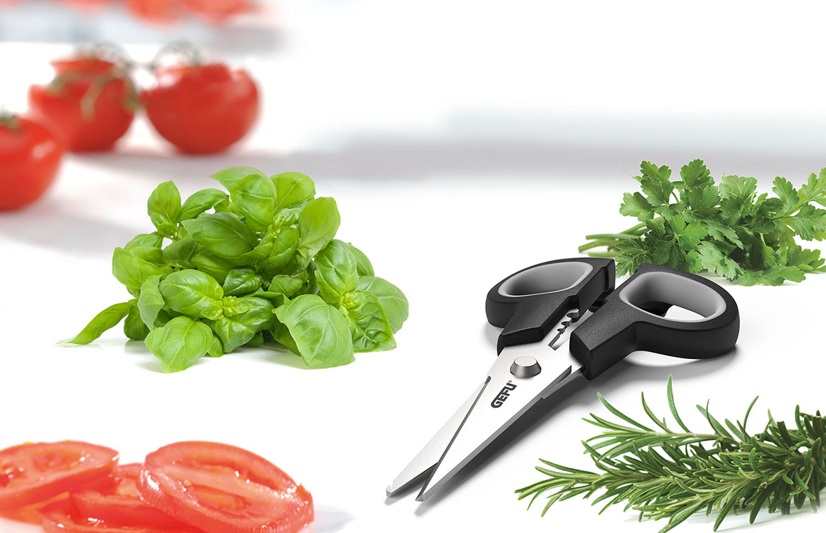 Multifunktions-Küchenschere mit Kräutern und Tomaten Eine schwarze Küchenschere neben frischen Kräutern wie Basilikum, Petersilie und Rosmarin, sowie geschnittenen Tomaten