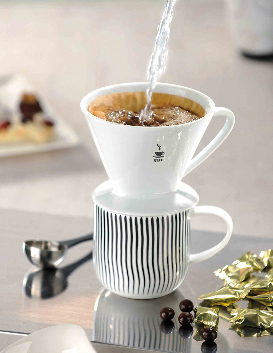 Ein weißer keramischer Kaffeefilter auf einer gestreiften Tasse. Heißes Wasser wird über den Kaffeefilter gegossen, um frisch gebrühten Kaffee herzustellen. Im Hintergrund sind Kaffeebohnen und verpackte Süßigkeiten zu sehen.