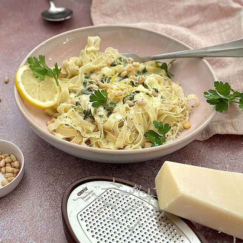 Leckere Tagliatelle mit Sahnesauce, Spinat und Parmesan Ein Teller mit Tagliatelle, garniert mit Sahnesauce, Spinat, Parmesan und einer Zitronenscheibe, dekoriert mit Petersilie und Pinienkernen.