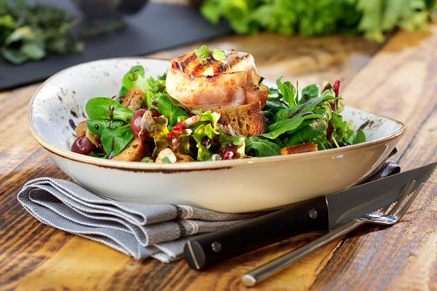 Brotsalat mit gegrilltem Ziegenkäse im Speckmantel Ein köstlicher Salat aus frischem Blattgemüse, gegrilltem Käse und Croutons, serviert in einer Schale auf einem Holztisch, daneben liegt Besteck.