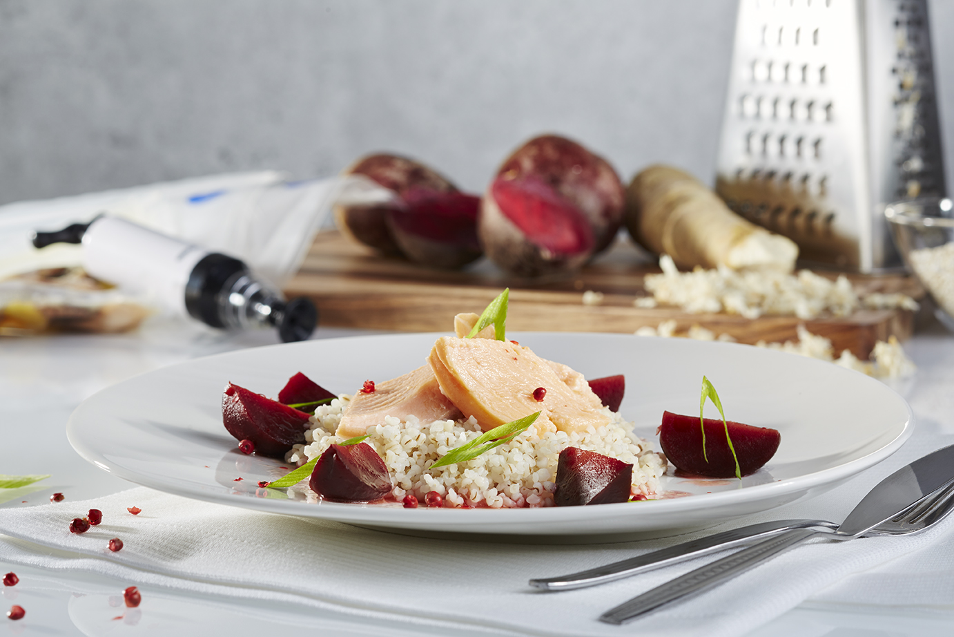 Sous vide Saibling mit Roter Bete, Graupen und Meerrettich Nahaufnahme eines Tellergerichts mit zartem Hähnchenfilet auf Bulgur, garniert mit Roter Bete und frischen Zwiebeln. Im Hintergrund sind eine Reibe und frische Zutaten zu sehen.