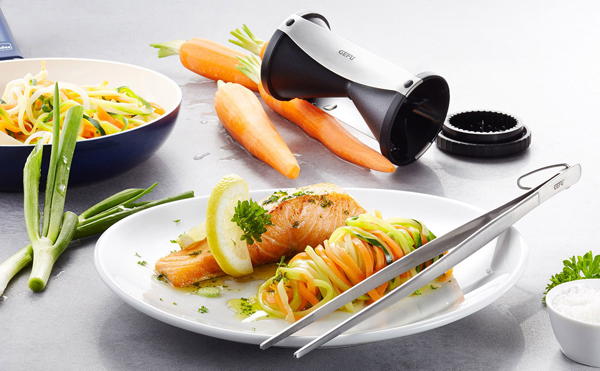 Zoodles mit Lachs und Zitronenscheibe auf weißem Teller Ein Gericht mit Zucchini-Nudeln, Karotten und Lachsfilet serviert mit Zitronenscheibe, neben einem Spiralschneider und Frühlingszwiebeln.