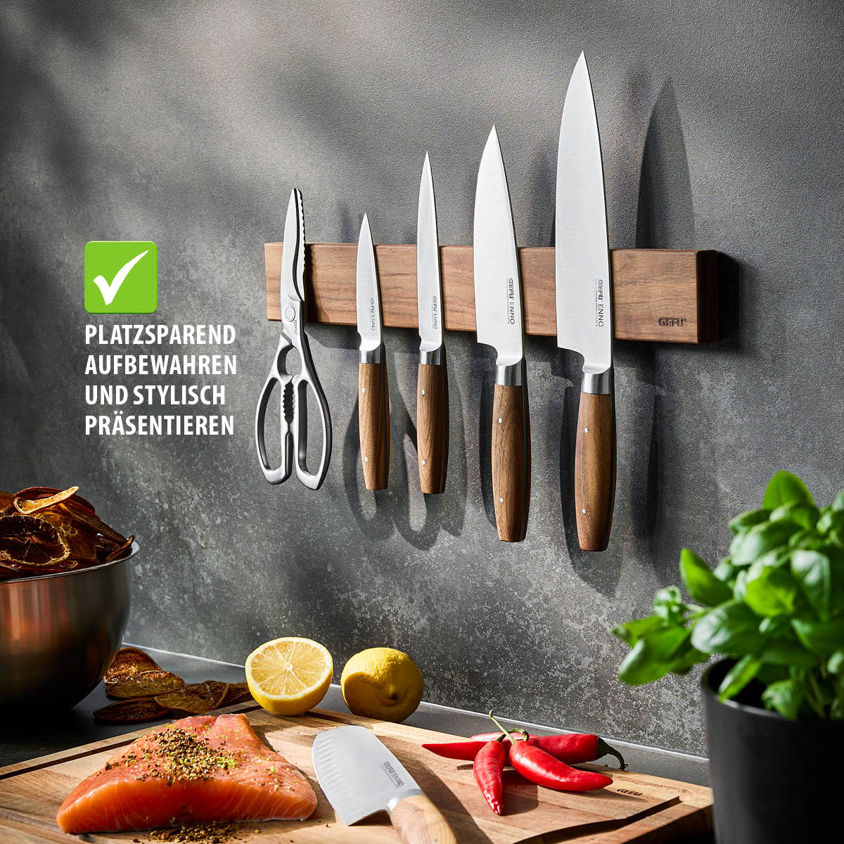 Eine magnetische Messerleiste aus Holz an der Wand, bestückt mit Küchenmessern und einer Schere. Darunter liegt frischer Lachs, Zitronen und Chili auf einem Schneidebrett.