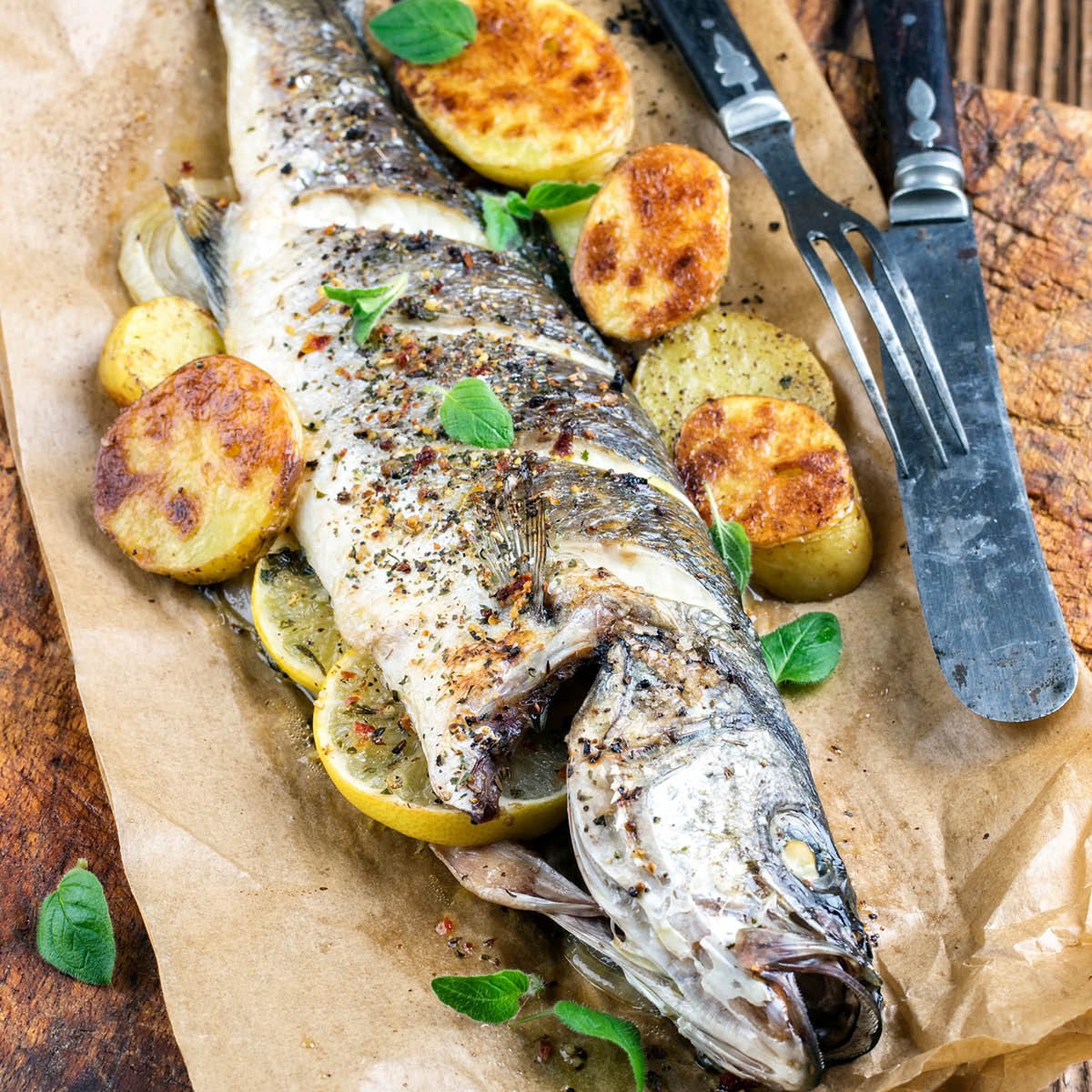 Ofenfrischer Fisch mit gebratenen Kartoffeln und Kräutern Ein ganzer, ofengebackener Fisch mit Zitronenscheiben und knusprigen Kartoffeln, garniert mit frischen Kräutern, serviert auf Backpapier.