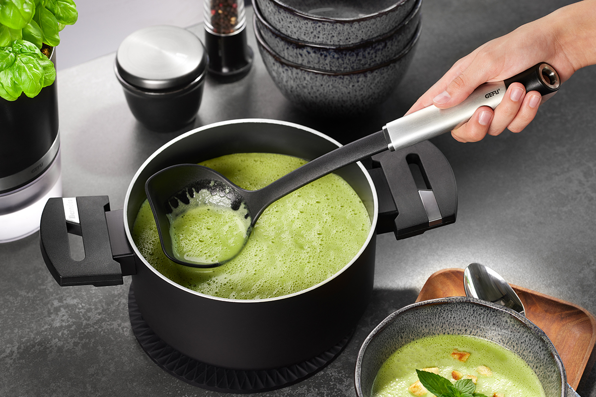 Kochtopf mit grüner Suppe und Kelle in moderner Küche Hand hält Kelle über einen Kochtopf mit grüner Suppe, daneben Schalen und Gewürzmühlen auf grauem Tisch