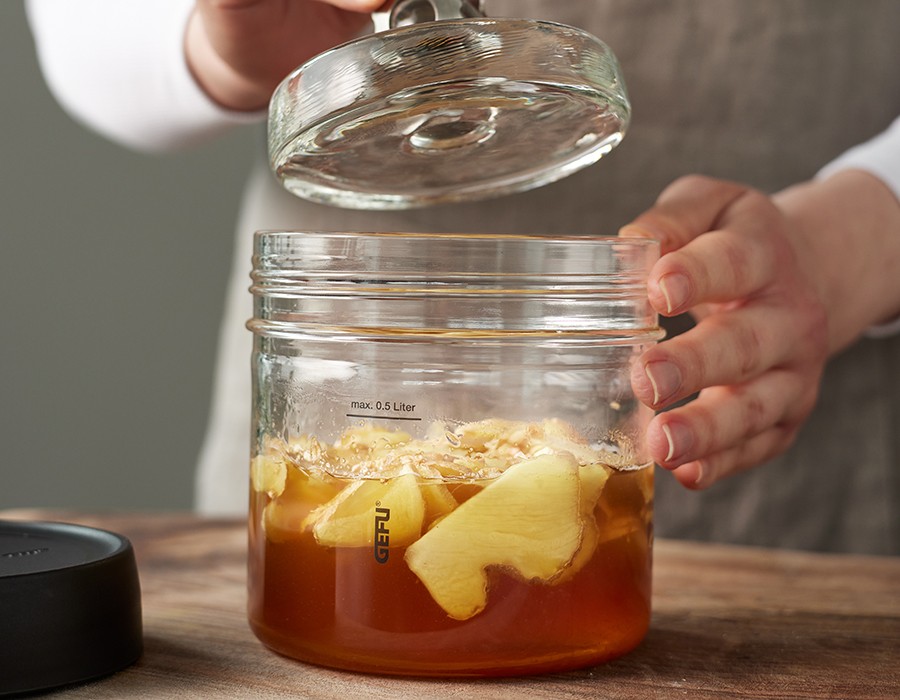Ginger Bug  Person öffnet Glas mit Ingwerstücken und Flüssigkeit zur Fermentation