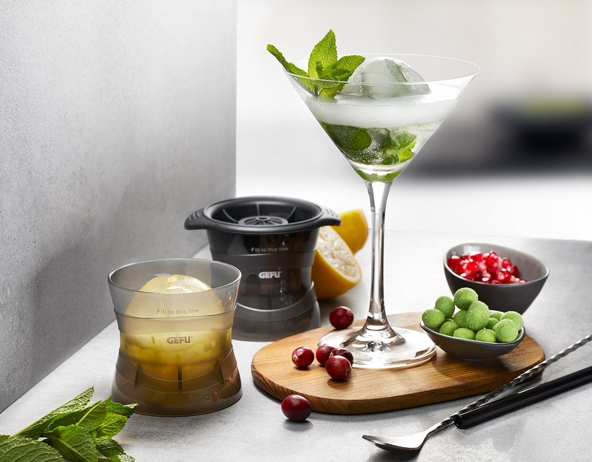 Cocktailglas mit Eisball und Minze neben Eiskugelformer Ein elegantes Cocktailglas mit einem Eisball und Minzblättern, flankiert von einem Eiskugelformer, einer Zitronenhälfte und Schalen mit Obst und grünen Süßigkeiten.