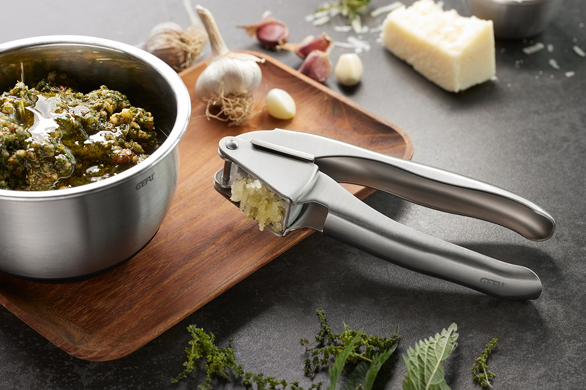 Edelstahl-Knoblauchpresse auf Holzbretter mit frischem Knoblauch, Pesto in Edelstahlschüssel und Parmesanstück im Hintergrund