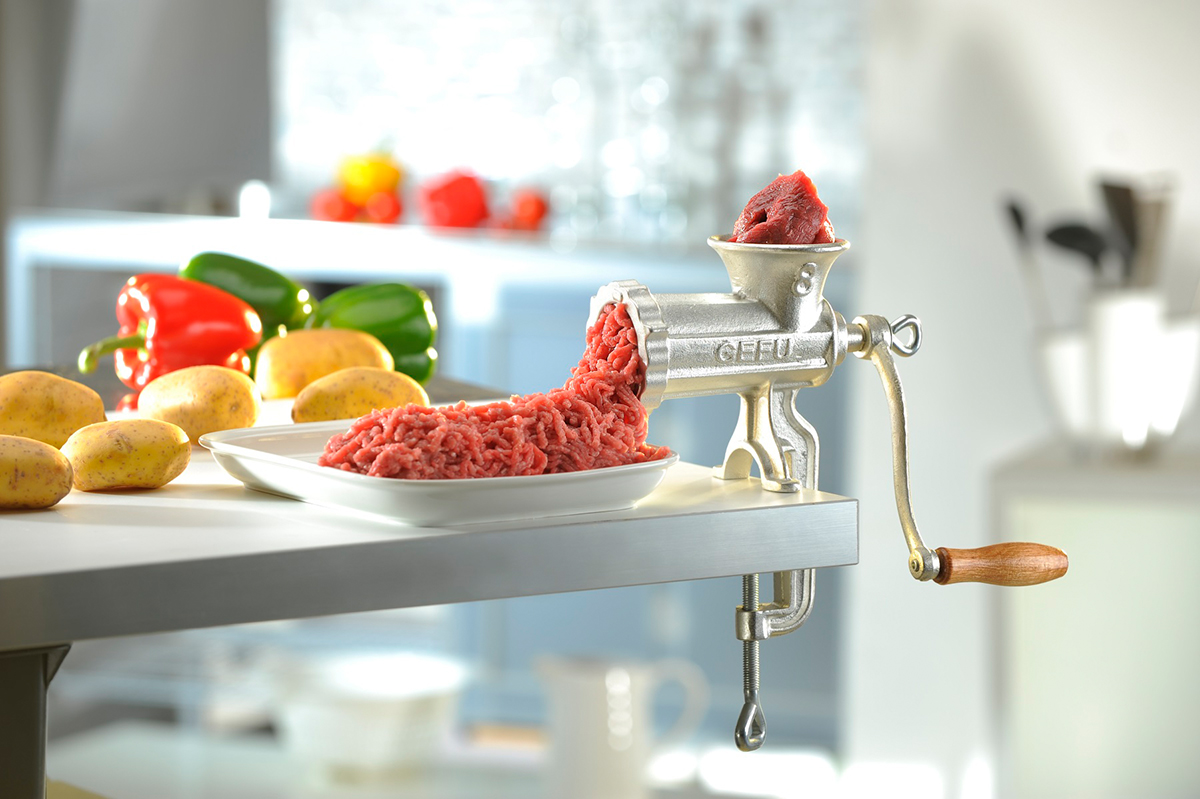 Manuelle Fleischwolf-Maschine aus Metall in moderner Küche Ein manueller Fleischwolf aus Metall klemmt an einem Küchentisch und verarbeitet Fleisch zu Hackfleisch. Daneben liegen Kartoffeln und Paprika.
