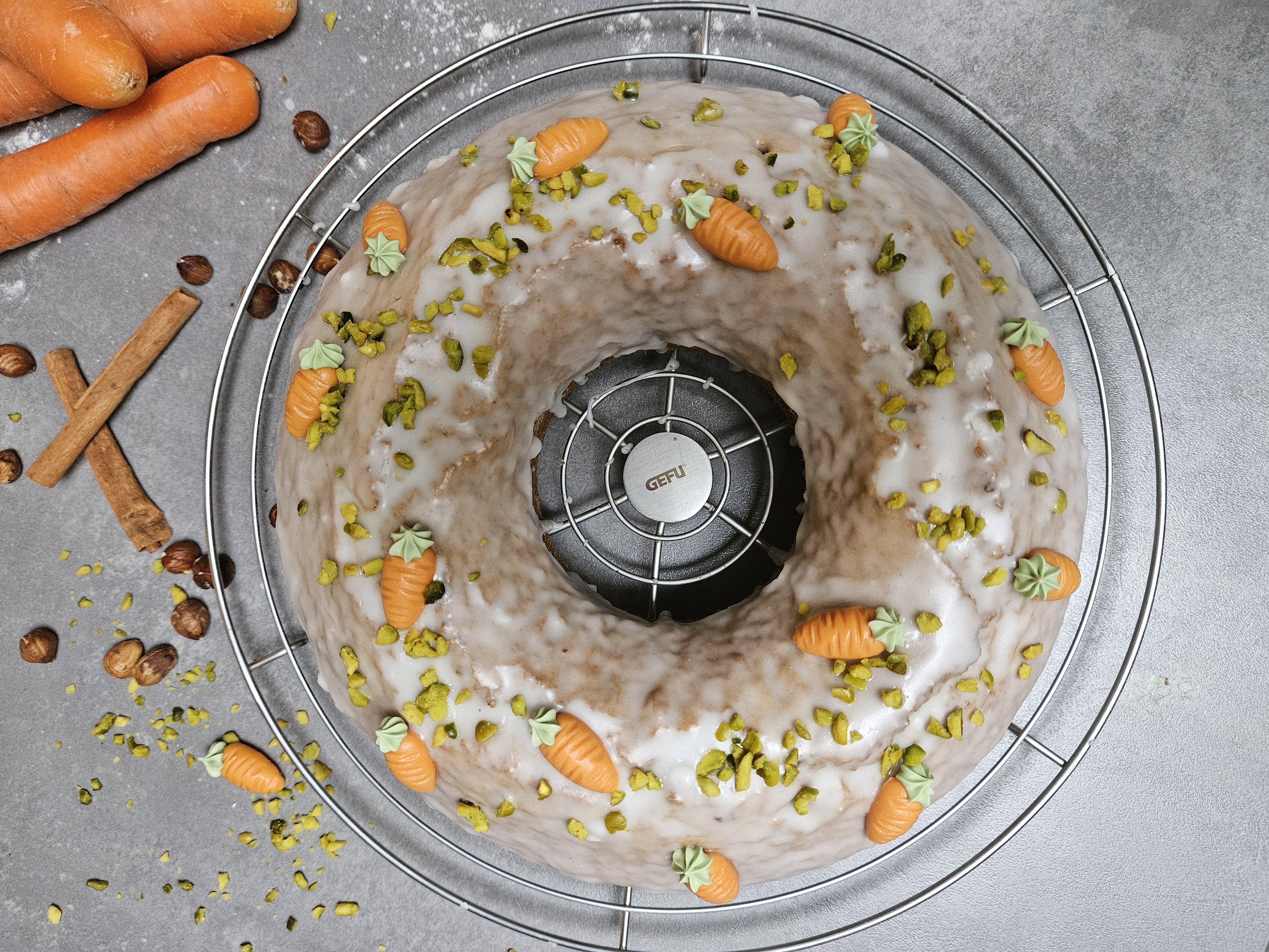 Rüblikuchen   Ein Ring-Karottenkuchen bedeckt mit Zuckerguss und dekoriert mit Marzipankarotten und gehackten Pistazien, umgeben von Haselnüssen, Zimtstangen und Karotten.