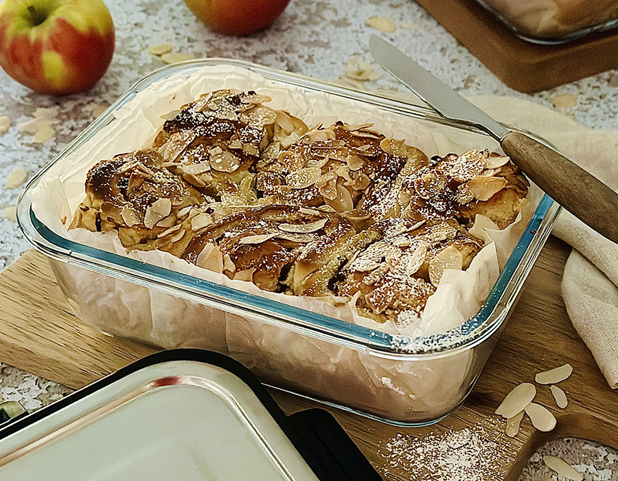 Apfel-Mandel-Schnecken Ein köstlicher Apfel-Blechkuchen bestreut mit Mandeln, serviert in einer transparenten Glasform. Im Hintergrund sind frische Äpfel und ein Schneidemesser zu sehen.