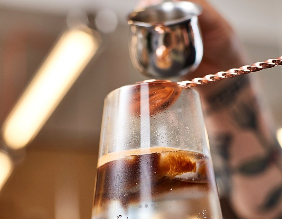 Espresso Tonic Glas mit Eiswürfeln, in das ein Barista kalten Kaffee aus einem kleinen Kännchen gießt