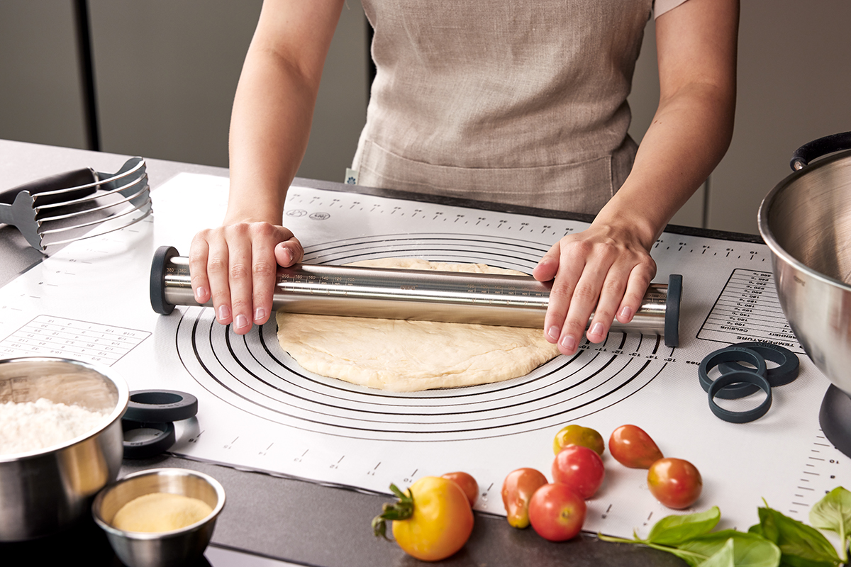 Teig ausrollen auf einer Backmatte mit Edelstahl-Teigrolle Eine Person rollt Teig auf einer Küchenmatte mit einer Edelstahl-Teigrolle aus, umgeben von Zutaten wie Mehl und Tomaten.