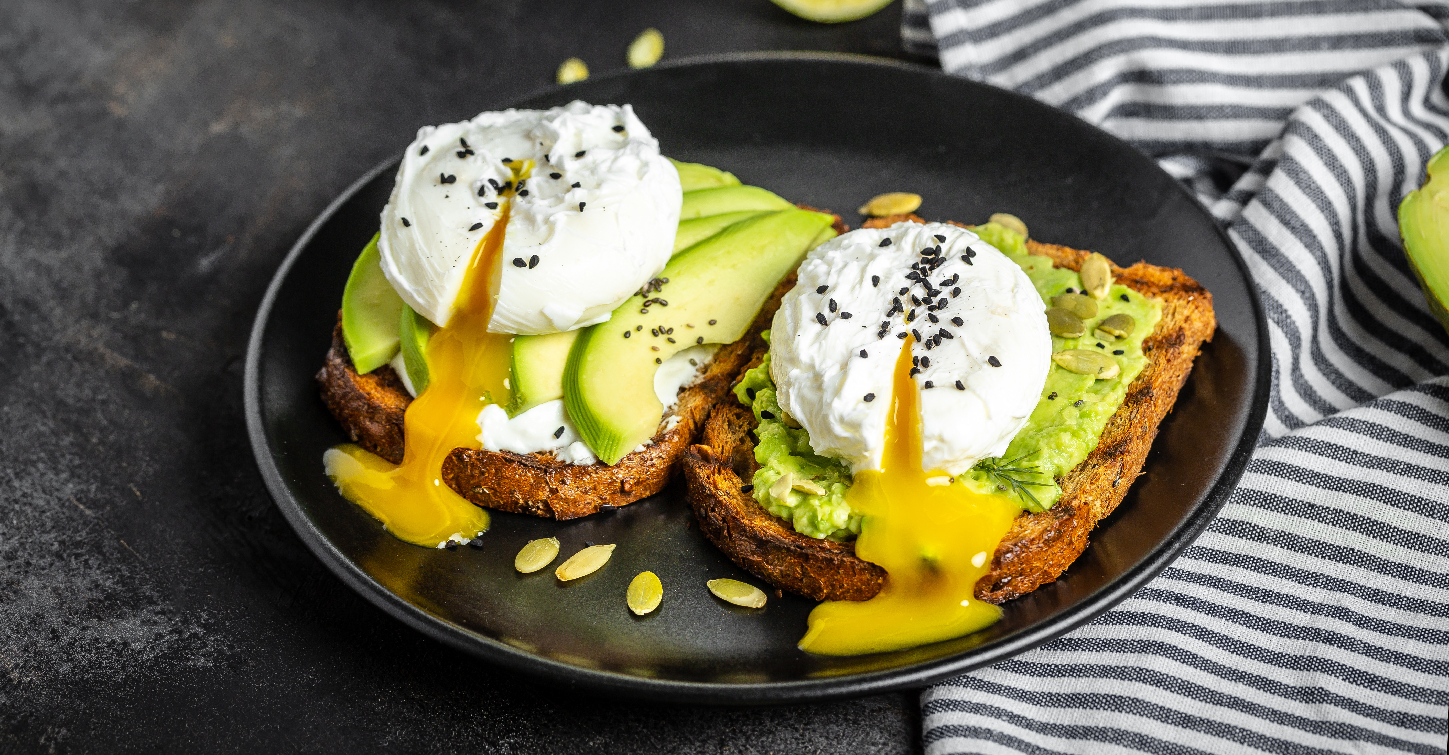 Avocado-Toast mit pochierten Eiern  Zwei Scheiben Avocado-Toast mit pochierten Eiern und schwarzem Sesam, garniert mit Kürbiskernen, auf einem schwarzen Teller serviert