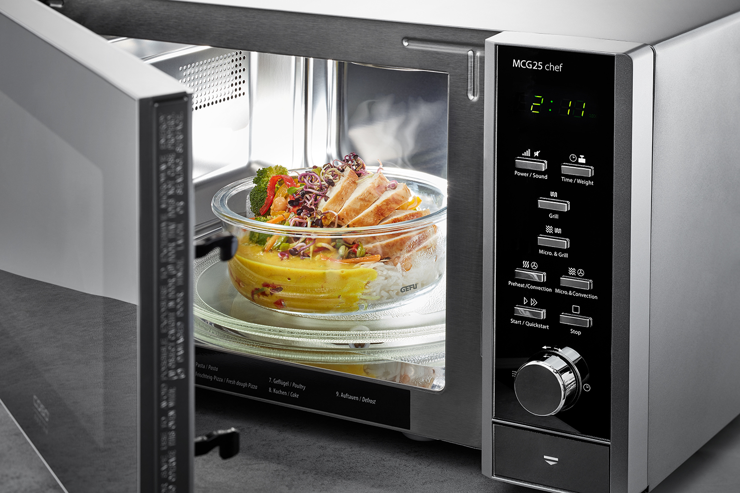 Multifunktions-Mikrowelle mit Glasbehälter und Mahlzeit Eine geöffnete Mikrowelle zeigt ein Gericht mit Hähnchen, Reis und Gemüse in einem Glasbehälter.