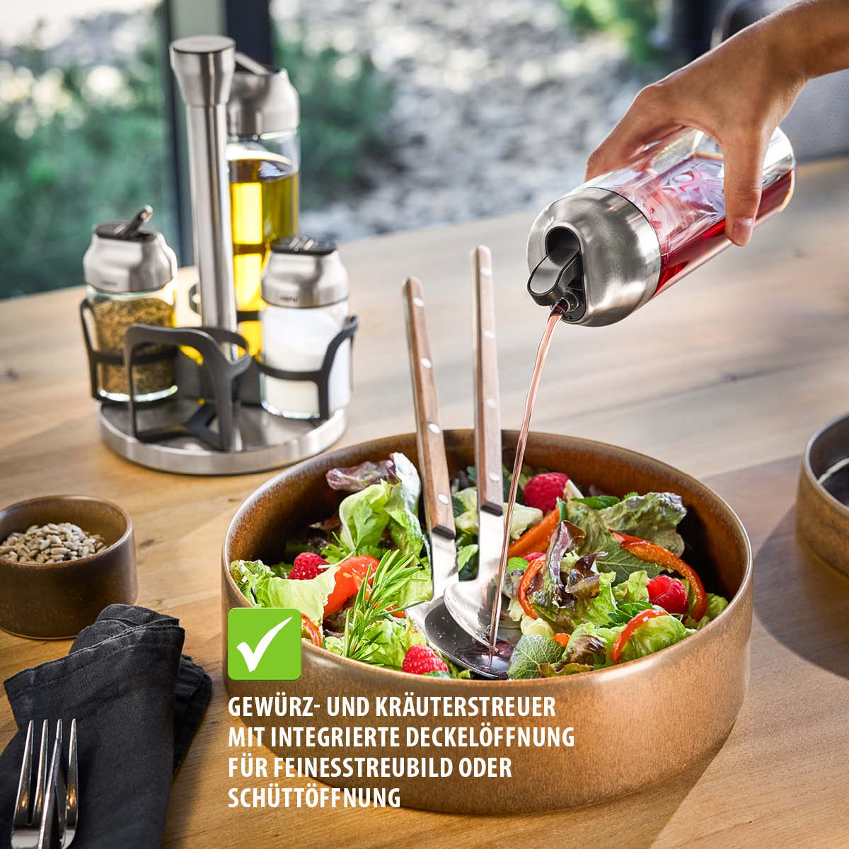Eine Hand gießt Sauce aus einem Gewürzstreuer über einen frischen Salat, mit einer Auswahl an Ölen und Gewürzen im Hintergrund.