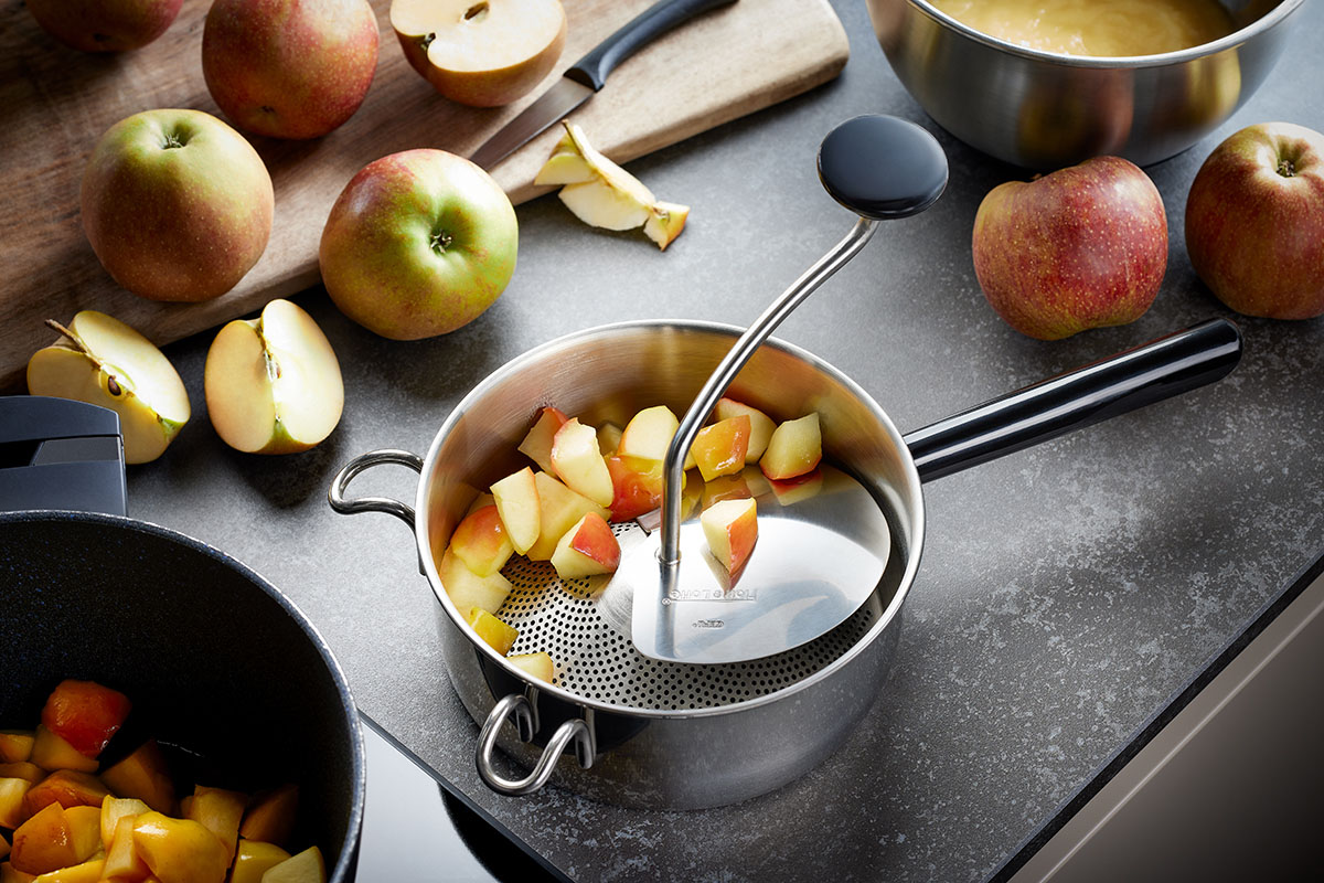 Moderne Küchenarbeit mit Passiergerät und frischen Äpfeln Ein Passiergerät mit Apfelstücken, umgeben von frischen Äpfeln auf einem Schneidebrett in einer modernen Küche.