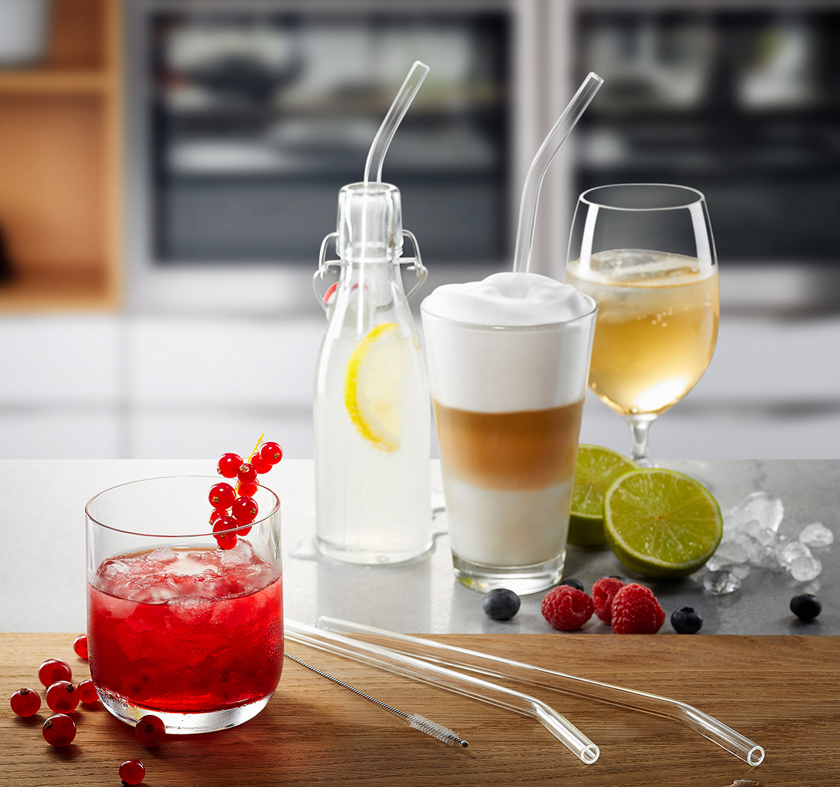 Vier verschiedene Getränke mit Glasstrohhalmen auf einem Tisch, darunter ein roter Cocktail, ein Latte Macchiato, ein Zitronenwasser und ein Weißweinglas, umgeben von Limettenscheiben, Beeren und Eiswürfeln.
