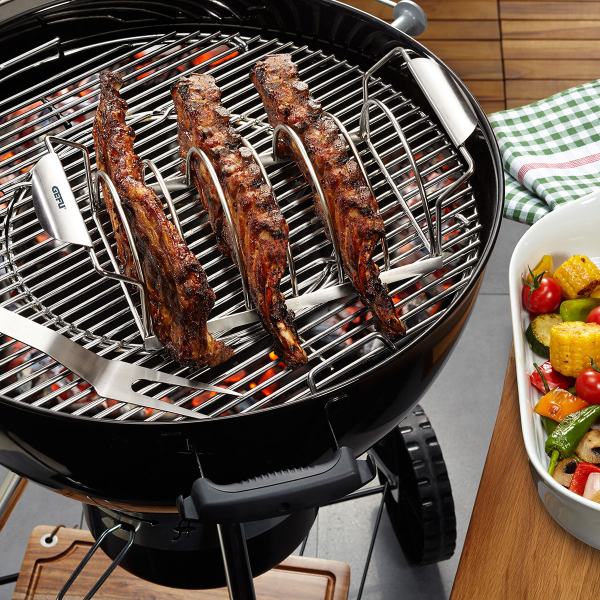 Holzkohlegrill mit gegrillten Rippchen und Gemüsebeilage Ein Holzkohlegrill mit drei gegrillten Rippchen in einem Grillständer und einer Schale mit gegrilltem Gemüse auf einem Tisch