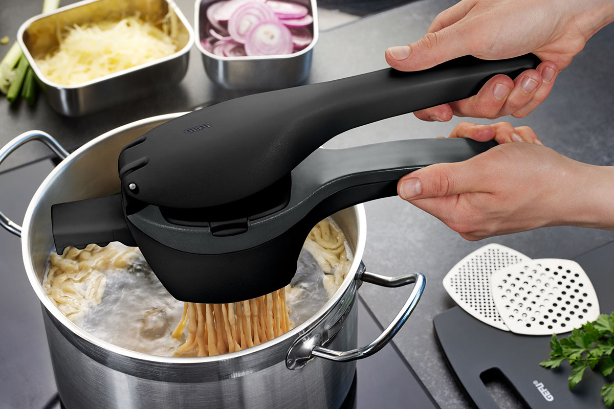 Eine Person benutzt eine Spätzlepresse, um frische Spätzle in einen kochenden Topf zu drücken. Im Hintergrund sind geschnittene Zwiebeln und geriebener Käse zu sehen.